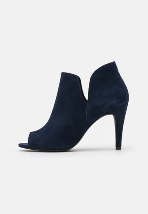 Escarpin bleu marine en daim à bout ouvert avec une silhouette courbée et des côtés découpés. Présente une semelle noire élégante et une quincaillerie minimale.