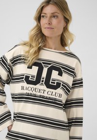 Gestreiftes langärmliges Oberteil in Beige mit schwarzen horizontalen Streifen. Verfügt über das "JC"-Logo und den Text "RACQUET CLUB" in einem kontrastierenden Design.