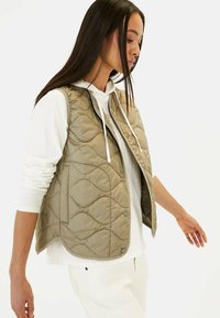 Beige gesteppte Weste mit hohem Kragen, Druckknöpfen und einer glatten Textur. Getragen über einem weißen Hoodie und lockeren weißen Hosen.