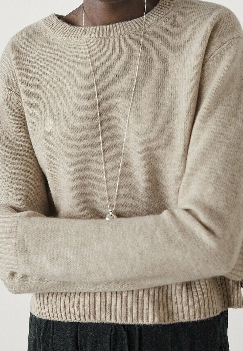 Personne portant un pull en maille beige avec les manches longues retroussées, et un fin collier en argent avec un pendentif rond.