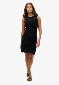 Robe noire sans manches avec des motifs en tricot texturé, un ourlet en forme de coquille et une silhouette ajustée. Portée avec des sandales plates noires.