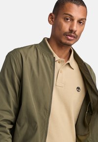 Giacca bomber verde oliva realizzata in tessuto morbido con colletto a coste, indossata sopra una polo beige chiaro con logo ricamato.