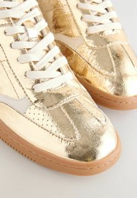 Goldene Metallic-Sneaker mit weißen Schnürsenkeln, Lederobermaterial, perforierten Details und brauner Gummisohle. Sie zeichnen sich durch ein klassisches sportliches Design aus.