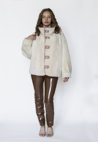 Krämfärgad jacka i fejkpäls med rosa accentknappar, hög krage och vida ärmar, kombinerad med glansiga bruna vinylbyxor och nakna öppna mules.