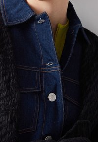 Mörk denimskjorta med orange sömmar, har silvriga knappar och en krage. Under finns ett strukturerat grönt plagg. Svart tyg är också synligt.