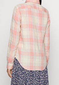 Camisa de botones en patrón de cuadros rosa y crema, tejido ligero, dobladillo curvado, mangas largas con cierres de botones, llevada sobre una falda floral.