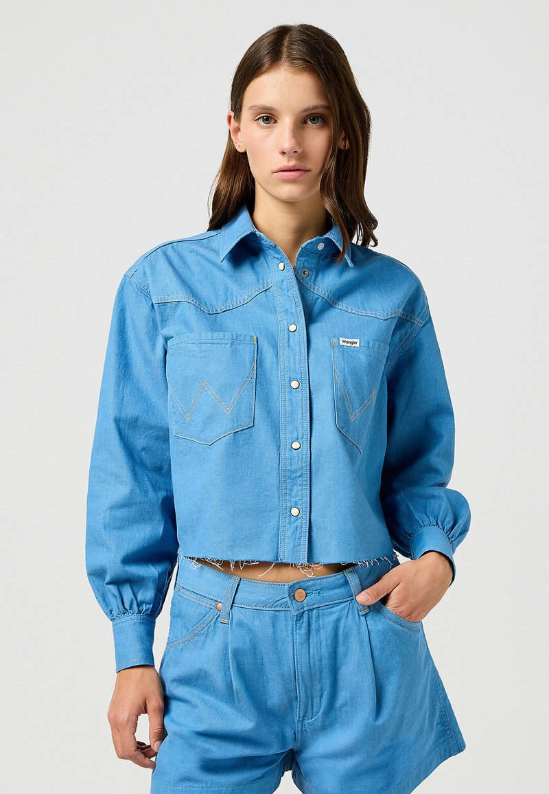 Ljust blå cropped denimskjorta med knäppningar, två bröstfickor och subtila sömmar. Långa puffärmar framhäver designen.
