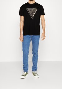 Mann trägt ein schwarzes Guess-T-Shirt mit Logo, blaue Jeans und olivgrüne Sneakers und steht vor einem schlichten weißen Hintergrund.