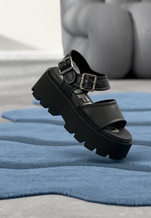 Black chunky platform sandal with two silver buckles on blue textured rug, blurred gray sculpture in background.
