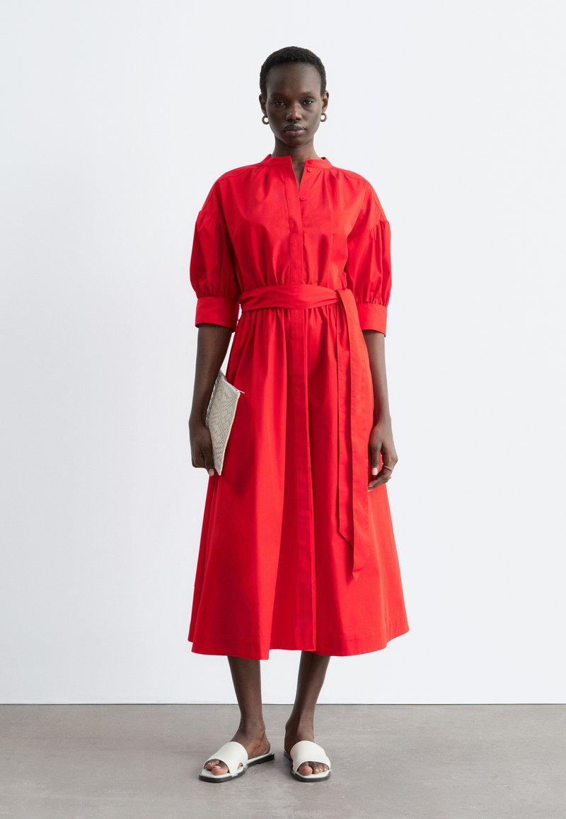 Femme portant une robe midi rouge à manches bouffantes et ceinture à la taille, tenant une pochette beige texturée, et chaussée de sandales blanches à enfiler.