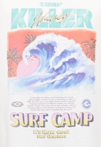 Valkoinen t-paita, jossa on värikäs grafiikka aallosta ja palmuista sekä tekstit "SURF KILLER" ja "SURF CAMP" eri paksuuksilla.
