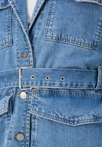 Giacca in denim azzurro chiaro con vita regolabile, due tasche frontali, accessori in metallo argentato e dettagli di cucitura gialla a contrasto.