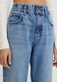 Jeans de mezclilla azul claro con una cinturilla fruncida, estilo clásico de cinco bolsillos y sutiles detalles de costura naranja en las costuras.