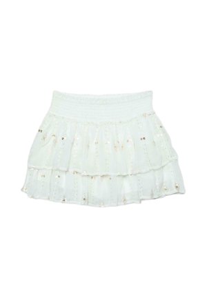 Frau mit nassen Haaren, die ein weißes Bikinioberteil, einen mehrlagigen weißen Minirock und beige Sandalen trägt und vor einem schlichten braunen Hintergrund steht.