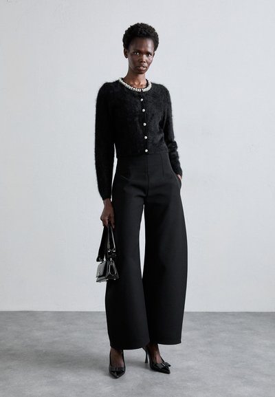 Schwarze, kurz geschnittene Strickjacke mit Perlenknöpfen, kombiniert mit hoch taillierten, weiten schwarzen Hosen. Das Model hält eine kleine schwarze Handtasche und trägt schwarze Absätze.