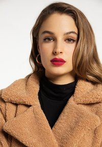 Beige faux fur coat with a notched collar, displayed over a black turtleneck. Model has a bold red lip and large hoop earrings.
