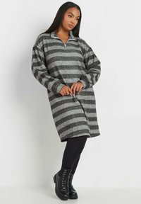 Abito a pullover a strisce grigie e nere con colletto zip, maniche lunghe e vestibilità ampia. Indossato con leggings neri e stivaletti con lacci.