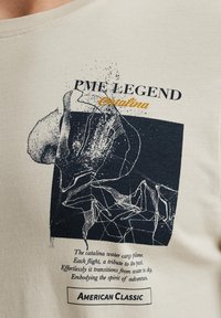 Krémové tričko s grafikou v námořnické modři a oranžovými akcenty. Text zahrnuje "PME LEGEND Catalina" a "AMERICAN CLASSIC."