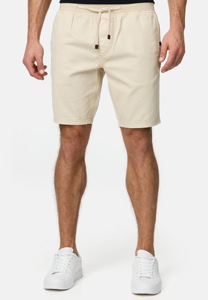 Homme portant un short beige à cordon et des baskets blanches, debout devant un fond clair uni.