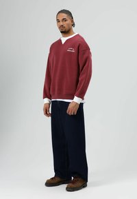 Bordeauxrode sweatshirt met een wit logo, ontspannen pasvorm, geribbelde manchetten over een wit shirt, donkerblauwe wijde jeans en bruine wandelschoenen.