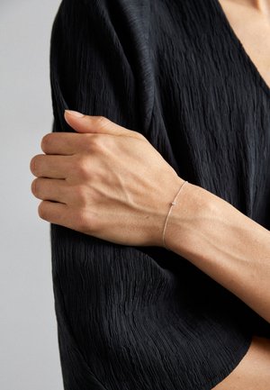 Silver bracelet with a delicate chain and small stone detail, worn on a lightly tanned hand, against a textured black fabric background.