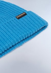 Stickad mössa i klarblått, med ribbad textur och vikt kant. En liten svart etikett med vit text syns på sidan.