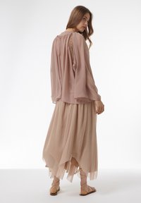Bluse in Blush mit langen Glockenärmeln; offene Rückseite mit Spitzenverzierungen. Geschichteter, fließender, knöchellanger Rock in einem passenden hellen Farbton.