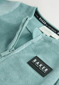 Indumento in pile verde acqua chiaro con chiusura a mezza zip. Presenta un'etichetta nera con la scritta "BAKER by TED BAKER". Morbida texture in tutto l'indumento.