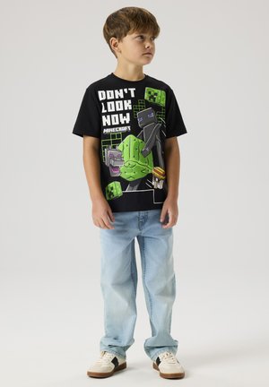 Dreng står og kigger til siden iført en sort Minecraft-tema T-shirt med figurer og tekst, lyseblå jeans og hvide sneakers.