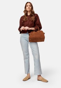 Woman wearing brown buttoned blouse and light blue jeans holding a large brown suede handbag, beige slip-on shoes, standing on white background.