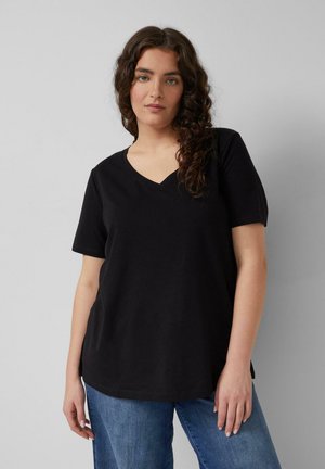 Jeune femme aux longs cheveux bouclés portant un T-shirt noir à col en V et un jean bleu, posant devant un fond gris clair uni.