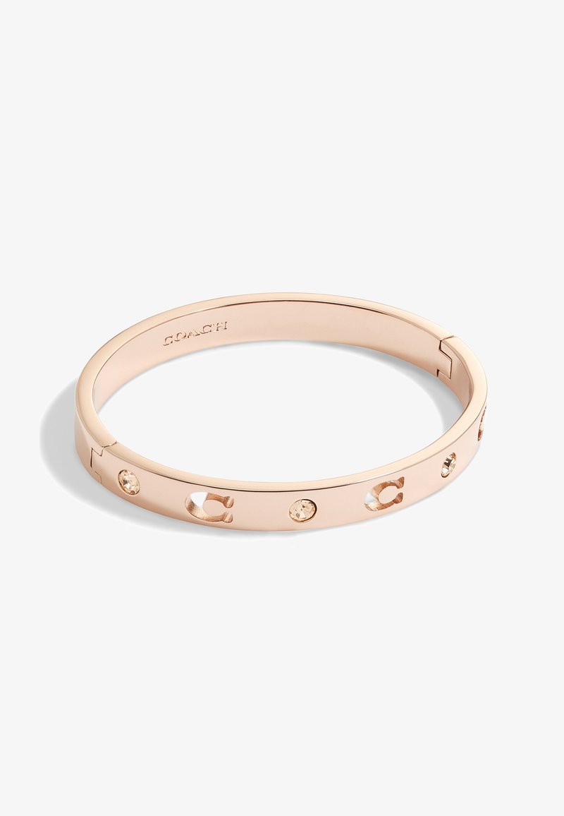 Bracelet jonc en métal rose doré gravé de la lettre "C" et orné de petits cristaux, avec le nom de la marque "Coach" à l'intérieur du jonc.