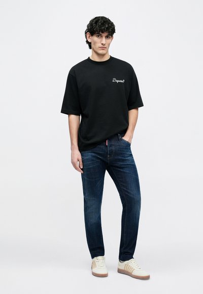 Melna kokvilnas T-krekls ar logotipu uz krūtīm, kombinēts ar tumši zilām džinsa biksēm. Modelis valkā baltas sporta kurpes ar brūnganiem akcentiem. Vienkārša stilizēšana.