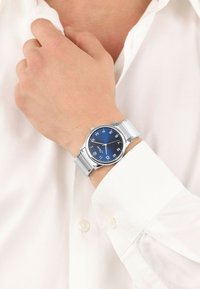 Silberne Armbanduhr mit blauem Zifferblatt und weißen Ziffern, ausgestattet mit einem Metallarmband und einem runden Gehäuse, getragen an einer Hand, die gegen ein weißes Hemd gehalten wird.