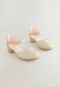 Par af guldglitrende lavhælede sko med ankelremme og dekorative spænder, på en neutral baggrund.