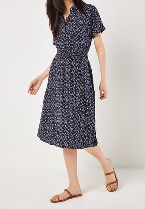 Femme portant une robe bleu marine à motifs jusqu'aux genoux avec des manches courtes et des sandales plates marron marchant sur un fond blanc.