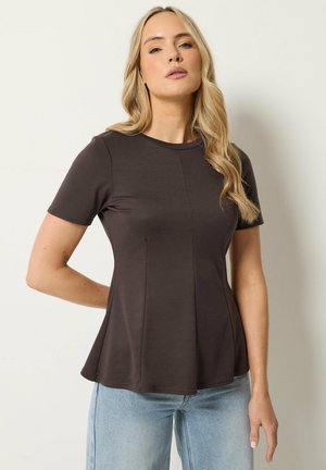 Lyshåret kvinde med løse bølger iført en tætsiddende kortærmet mørkebrun top og lyseblå jeans, stående mod en ensfarvet baggrund.