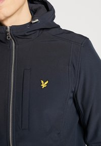 Marineblauwe capuchonjas met een ritssluiting, voorzien van een borstzak en een geborduurd geel vogellogo aan de linkerkant. De stof heeft een gladde textuur.