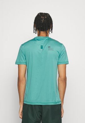 Mann mit geflochtenem Haar, der ein kurzärmliges Sportshirt von Lacoste in Türkis und dunkle Shorts trägt, von hinten vor einem weißen Hintergrund gezeigt.