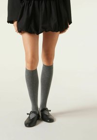 Schwarze Lederschuhe mit abgerundetem Zehenbereich, einem kleinen Schnallendetail, getragen mit dunkelgrauen knielangen Socken. Der Rock ist voluminös und schwarz.