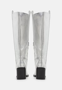 Bottes montantes argentées en matériau brillant, avec des talons carrés noirs et des fermetures éclair latérales pour un enfilage facile. Texture lisse, design minimaliste.