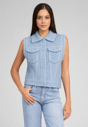 Femme aux longs cheveux foncés portant un gilet sans manches bleu clair boutonné et un jean bleu clair, debout sur un fond gris.