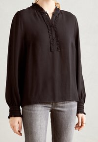 Blusa nera a maniche lunghe con sottili righe verticali, colletto arricciato, chiusura con bottoni e polsini arricciati, abbinata a jeans grigi.