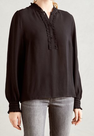 Schwarze Bluse mit langen Ärmeln, dezenten vertikalen Streifen, Rüsche am Kragen, Knopfschluss und gerafften Manschetten, kombiniert mit grauen Jeans.