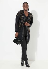 Kimono negro de cuentas con patrones geométricos, dobladillo con flecos, combinado con una blusa y pantalones negros, y un pequeño bolso negro.