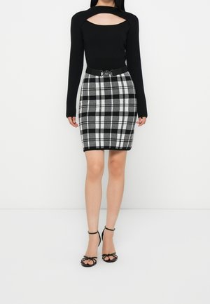 Woman wearing black long-sleeve top with chest cutout, black-and-white plaid skirt with belt, and black open-toe heels standing against white background.