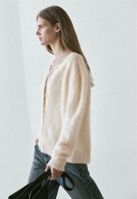 Light cream cable-knit cardigan with a relaxed fit, featuring a round neckline and subtle button details, paired with blue jeans and a black bag.