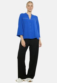 Woman wearing a royal blue buttoned blouse with lace detail, black wide-leg pants, and white sneakers, standing against a plain background.