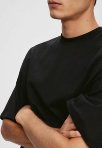 Joven con piel clara que lleva una camiseta negra holgada, con los brazos cruzados, mirando ligeramente hacia la derecha y con una expresión neutral.