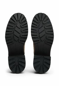 Une paire de chaussures à semelle en caoutchouc noire avec des motifs de semelle profonds, présentée de dessous sur un fond blanc.